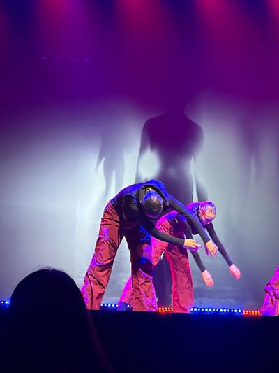 The image depicts a stage performance taking place in a venue within the city of Ventspils, Latvia. The scene is dominated by vibrant purple and magenta stage lighting, creating a dramatic atmosphere.

In the foreground, the top of an individual's head, likely an audience member, is visible and out of focus in the bottom left corner. The immediate stage edge features two rows of small LED lights: a segment of red lights on the left, a central segment of blue lights, and another segment of red lights on the right.

At least two performers are visible on stage, both adorned in long-sleeved dark tops and loose-fitting, shiny reddish-brown or maroon pants. Their heads are covered with reflective, textured caps or masks, possibly embellished with sequins or beads, which shimmer under the stage lights. Both performers are captured mid-motion, leaning forward at the waist with their arms extended downwards and slightly forward, palms open. Their posture suggests a synchronized and expressive movement. A third performer's lower legs and part of their pants are visible on the far right of the frame, indicating additional participants.

The background consists of a large white or light-colored screen or wall, onto which large, elongated silhouettes of human figures are projected or cast as shadows. These shadows are significantly larger than the visible performers and appear dark against the lighter, somewhat hazy background. The overall lighting scheme suggests a theatrical or dance production with intentional use of color and shadow play.