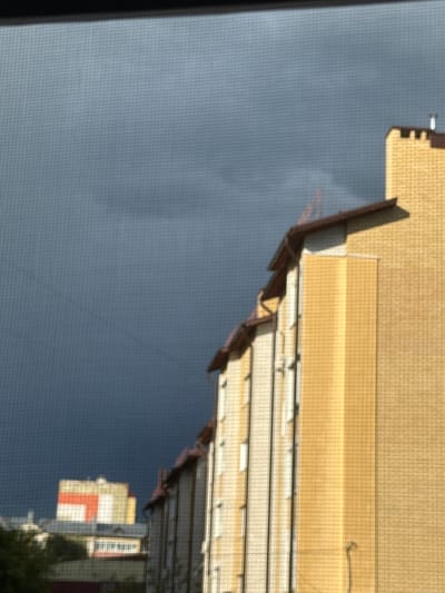 The image shows a view from a window with a screen, looking out at buildings against a dark, cloudy sky. The foreground is dominated by the screen pattern. In the mid-ground, the side of a large, multi-story building with yellow brickwork is visible, featuring cream-colored vertical elements and brown roof edges. Further in the background, another building with red and yellow sections is partially obscured by trees. The overall impression is of an urban or suburban landscape under heavy cloud cover. The location is identified as Morozovka, Russia.
