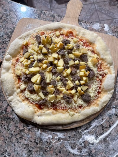 A circular, uncooked pizza rests on a light wooden pizza peel positioned on a speckled, polished granite countertop in a kitchen in Porto Alegre, Brazil. The pizza features a raw dough crust with a visible cornicione, covered with a layer of red tomato sauce. A substantial amount of shredded white cheese forms the base topping, over which numerous small, irregular cubes of a yellowish-brown cheese are scattered. Interspersed among the cheeses are dark brown, irregular pieces of cooked meat, likely beef. Traces of white flour are visible on the granite countertop to the lower right of the pizza, indicating recent food preparation. Artificial overhead lighting illuminates the scene.