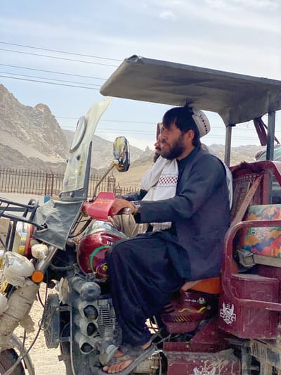This outdoor portrait shot captures a man operating a three-wheeled motorized rickshaw, possibly in Panjwai, Afghanistan, given the location context. The main subject, a man with a dark beard and dark hair, is seated on the driver's side, looking slightly to his right with a neutral expression. He wears a dark traditional garment, likely a shalwar kameez, a white patterned cap, and a white, intricately patterned scarf draped over his shoulder. A second man is partially visible behind him.

The vehicle is a modified motorcycle-based three-wheeler, or auto-rickshaw, with a reddish-brown body and a simple grey canopy providing shade. The handlebars, engine, and a worn windshield are visible. The driver is wearing open-toed sandals. In the background, arid, rocky mountains stretch across the horizon under a bright, cloudy sky, suggesting a dry, mountainous environment. Power lines are visible running horizontally across the mid-ground. A metal fence separates the immediate foreground from the distant landscape.

The time of day appears to be bright daylight, possibly mid-morning or afternoon. The vehicle shows signs of wear and personalized additions, such as various stickers and wrapped components. Visible text includes "BE" on the fuel tank and "94LH-200" on a sticker below the driver's seat, possibly a registration number or model identifier. There is also some stylized script, likely Arabic or Farsi, on the fuel tank, possibly decorative or a brand name. The overall scene depicts typical daily travel in a rural or semi-urban setting within this region.