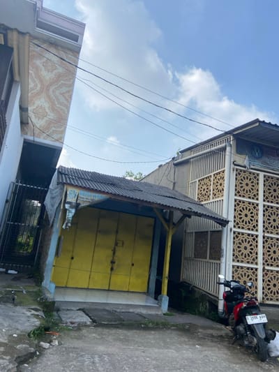 The image shows a narrow alleyway in Kecamatan Cimahi Selatan, Indonesia. The alleyway is lined with two buildings, one with a corrugated metal roof and the other with an intricate, carved wooden facade. The building with the corrugated roof has a yellow door. A motorcycle is parked beside the building with the wooden facade. The motorcycle has a license plate reading "D 3998 ABH." Overhead, a blue sky with white clouds can be seen. The alley is shaded by the buildings and a few trees. There are power lines running overhead. The pavement is made of concrete and is slightly uneven. There are some weeds growing along the sides of the alley.