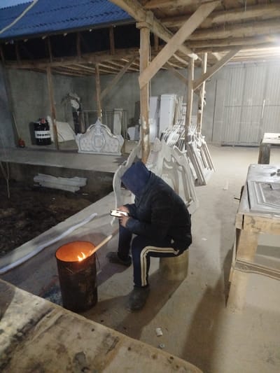 The scene depicts an interior view of what appears to be a workshop or storage facility in Andijan, Uzbekistan. The structure features exposed wooden support beams and a blue corrugated metal roof.
A single individual is observed in the foreground, seated on a cylindrical metal stool. The person wears a dark hooded jacket with the hood obscuring their face, dark trousers with a white stripe down the outer seam, and dark clogs. Their attention is directed downwards toward a smartphone held in both hands.
To the immediate left of the individual, a dark, rusty metal barrel contains an open fire, with visible flames and a wooden stick extending from the interior. The ground beneath the fire and the person is concrete, while further left, the ground transitions to exposed soil.
The background contains numerous white, ornate furniture components, appearing to be unfinished or recently cast pieces such as headboards and cabinet doors, stacked against walls and on the floor. A prominent ornate white item with gold accents, resembling a bed headboard, is visible on a raised concrete platform in the mid-left. A black barrel labeled "neomix" is also situated on this platform. A wooden workbench is partially visible on the far right. The environment is illuminated by artificial light sources.