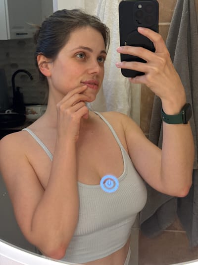 A woman is taking a selfie in a bathroom mirror. She is wearing a light grey tank top and an Apple watch with a green band. She is holding a black iPhone in her left hand. The background shows a tiled wall, a towel, and bathroom fixtures. The location in Kőröshegy, Hungary is not discernible from this image.