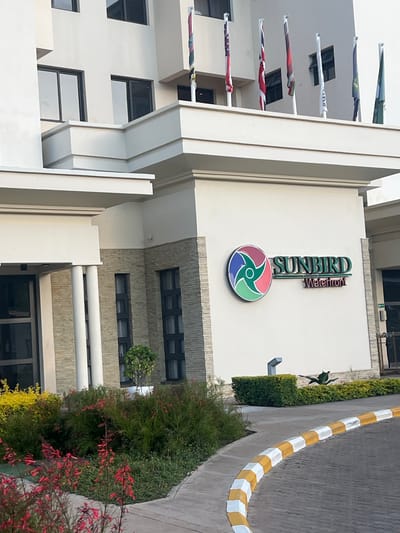 The image shows the Sunbird Waterfront hotel in Lilongwe, Malawi. The sign "SUNBIRD Waterfront" is prominently displayed on the exterior of the building above the entrance. Flags are visible on the left side of the building. A sidewalk with yellow and white curbs runs along the right of the image, with various plants and greenery visible in the foreground.