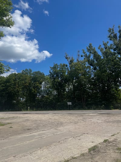The image captures a roadside scene in Konopnytsia, Ukraine. In the foreground, a rough, unpaved area with patches of concrete and dirt dominates the lower portion of the frame. A paved road runs horizontally across the middle ground. Beyond the road, a dense line of trees forms a natural barrier. The sky above is a vibrant blue, with scattered white clouds adding texture. A white sign with text in Ukrainian and English, "Зимна Вода / Zymna Voda," is visible to the left of the trees, indicating a nearby location. The overall environment suggests a rural or semi-rural setting. No people or vehicles are present in the image.