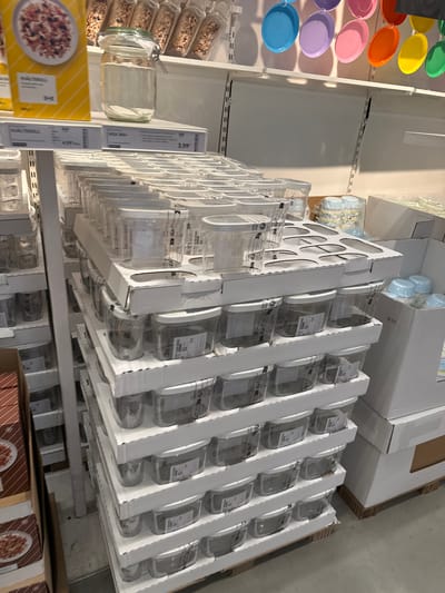This media file shows a retail display of glassware and food packaging in a store, likely a home goods store or supermarket. The primary subjects are rows of clear glass jars and containers, many filled with dry goods such as grains or granola. These are stacked neatly on shelves, some in their original packaging, suggesting they are for sale.
In the foreground and to the left, a selection of colorful plastic plates or bowls are displayed, in shades of green, yellow, orange, pink, purple, and blue. Further back, stacks of white boxes, some open to reveal more glassware, are visible. One of these boxes has a cut-out pattern on the side. On the right side of the image, more glassware is presented in white cardboard packaging, with multiple rows of identical containers.
Several price tags are visible. One clearly shows "3,99€" and "IKEA 365+". Another price tag reads "4,99€" and "HÜALTERROLL". The presence of IKEA branding on the price tag strongly suggests the location is an IKEA store. The lighting suggests an indoor setting with artificial lights, and there are no indicators of time of day or weather. The scene is clean and organized, typical of a retail environment. The overall impression is one of a well-stocked store shelf with a variety of products for home use, particularly kitchenware and food storage solutions.