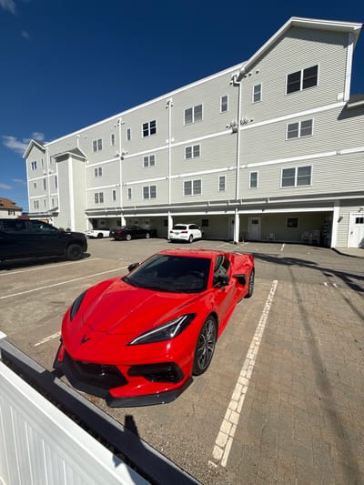 A bright red Chevrolet Corvette C8 is parked in a paved lot in front of a pale green, multi-story building. The building has a horizontal siding design and numerous windows. The Corvette is in the foreground, angled towards the right side of the frame, with its front end closest to the viewer. To the left of the Corvette, a black pickup truck is partially visible. Further back, other cars including a white Chevrolet Corvette and a dark sedan are parked. The ground is a light-colored brick paver surface with white painted parking lines. A white fence runs along the bottom left of the image. The sky is a clear, deep blue, with a few small white clouds visible towards the left. The scene appears to be a residential or commercial complex located in Hampton, United States of America, likely during daylight hours given the strong shadows.