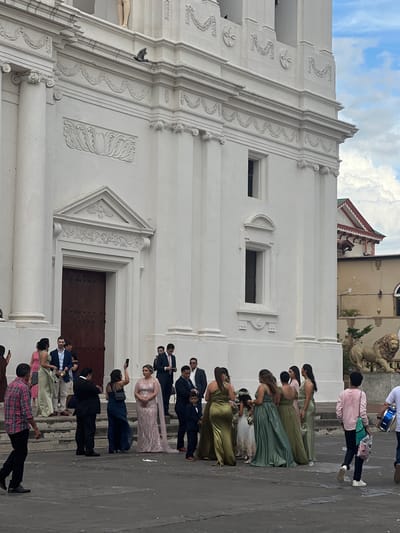 This image depicts an outdoor scene in front of a large, white, ornate building, likely a cathedral or historic church, located in León, Nicaragua. The architecture features classical columns, intricate carvings, and a large wooden double door. A group of people, dressed in formal attire, are gathered on the steps and the paved area in front of the building. Several individuals are holding smartphones, suggesting they are taking pictures or videos of an event, possibly a wedding or special ceremony.

In the foreground, a man in a plaid shirt walks away from the camera. To his left, a group of women in formal dresses, some in shades of green and pink, are standing together. Some of these women appear to be bridesmaids, and there are also young girls in white dresses, possibly flower girls. To the right of this group, a man in a suit is visible, along with other guests.

In the background, behind the main building, another building with a red and white decorative roofline is visible, along with a statue of a lion. The sky above is partly cloudy. The overall atmosphere suggests a social gathering or event taking place at a prominent landmark.