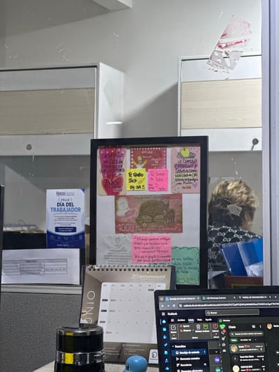 The image shows a desk with a bulletin board, a calendar, and a laptop. The bulletin board has several colorful notes and a Costa Rican 1000 colones banknote. A person with blond hair is partially visible in the background, seated at the desk, looking down at their laptop. The laptop screen displays an email interface and a WhatsApp chat window. The environment appears to be an office. The location is not discernible from the image provided.