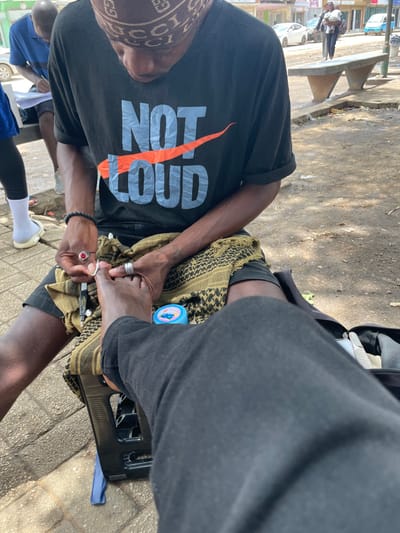 This media shows an outdoor scene in Luanda, Angola, during the daytime. The central focus is on two individuals engaged in a pedicure service on a paved surface, likely a sidewalk or informal market area.
The person performing the pedicure is seated, wearing a black t-shirt with the text "NOT GOOD" prominently displayed, where "GOOD" is crossed out by a large orange checkmark resembling a stylized swoosh. They also wear a patterned bandana or headwrap featuring the "GUCCI" logo. Their left arm, with visible veins, rests on a patterned scarf draped over their lap. They are holding nail clippers and are actively working on the foot of the second individual. A ring is visible on their right hand and a beaded bracelet on their wrist.
The second individual, whose legs are visible, is receiving the pedicure. They are wearing dark pants, and their left foot rests on a black plastic stool or stand. A small blue container is placed near their foot on the patterned scarf.
In the background, other people are partially visible, including one person in a blue shirt and white socks. Cars and what appears to be part of a bench or structure can also be seen further down the street, suggesting an urban or semi-urban environment. The bright natural light indicates it is daytime, with clear visibility and no signs of adverse weather. The overall impression is an informal street-side service.