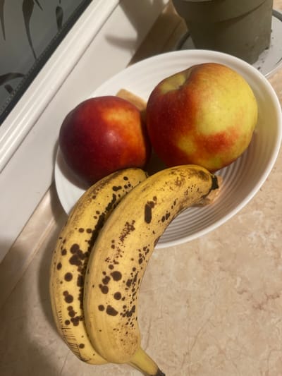 A close-up, overhead shot shows a white bowl filled with two apples and two bananas. The apples are red and yellow, and the bananas are yellow with brown spots. The bowl sits on a countertop that appears to be made of beige marble. In the background, a white ledge or counter is visible, and behind that, a dark object with a reflective surface. To the right of the bowl, a dark green potted plant sits on a white saucer. The scene is indoors, and the lighting suggests it could be a kitchen or dining area. The image does not contain any information that would allow for a determination of its location in Konstantynów Łódzki, Poland.