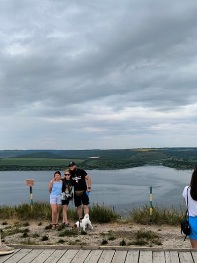 This image depicts an outdoor scenic overlook in the area of Horaivka, Ukraine, under an overcast sky. The foreground features a wooden platform transitioning to a dirt and sparse grass area where three individuals and a small dog are positioned. On the left, a brown information sign is mounted on a yellow and green striped pole. From left to right in the central group: a pre-teen female in a light blue tank top and white shorts stands beside a young adult female wearing sunglasses, a dark t-shirt, and dark shorts, carrying a dark shoulder bag. To their right, an adult male with a beard and a dark baseball cap wears a dark t-shirt, dark shorts, and a light-colored fanny pack; his left arm is around the central female's shoulders, and he holds a blue object in his right hand. A small, white and brown Jack Russell Terrier-type dog stands at their feet, appearing to be on a leash. A fourth individual, primarily visible from the back in a white t-shirt and blue shorts, is partially cropped on the far right. The midground is dominated by a wide, calm body of water. The background reveals undulating green hills and forested land, with interspersed patches of lighter green and yellow agricultural fields. The upper third of the image shows a sky completely covered by dense grey clouds.