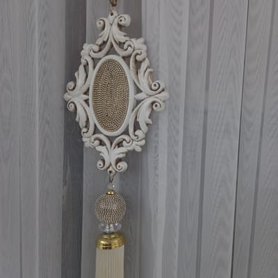 The image shows a decorative tassel hanging in front of sheer white curtains. The tassel has an ornate white frame at the top, filled with gold beads, from which hangs a crystal bead, a gold cap, and then a thick fringe of cream-colored threads. The curtains are vertically pleated and semi-transparent, revealing faint vertical stripes behind them. The scene is indoors, with no specific indication of its location within Briceni, Moldova.