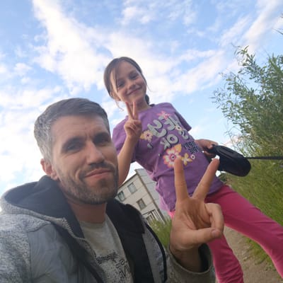 The image is a low-angle, wide-shot selfie featuring a man and a young girl against a sky with scattered clouds. The man, in the foreground on the left, is wearing a grey hooded jacket over a grey t-shirt, with grey hair and a beard. He is looking at the camera and has his right hand raised, displaying two fingers in a peace sign. The girl, positioned behind and to the right of the man, is wearing a purple t-shirt with floral designs and text, and bright pink pants. She is also looking at the camera and has her left hand raised in a peace sign. In the background, there is a multi-story building with several windows and a narrow pathway, suggesting an urban or semi-urban environment. Tall green grass and foliage are visible on the right side of the image. The overall setting appears to be outdoors. The girl is holding a black retractable dog leash in her right hand.