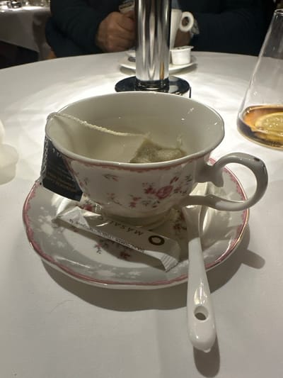 A teacup and saucer sit on a white surface. The teacup, adorned with a delicate pink floral pattern, contains a tea bag. A white spoon rests on the saucer. A packet of Masala tea sugar is also present. The scene is illuminated, casting shadows behind the cup and saucer. The image is taken from a high angle, capturing the arrangement with clarity.