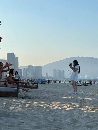The image captures a sunny day at My Khe Beach in Da Nang, Vietnam. In the foreground, several people are relaxing on beach chairs and loungers. To the left, a group of women are seated, with one woman holding up a phone to take a picture. Further to the right, a young woman in a white dress stands on the sand, holding a camera and appearing to take a photograph. In the background, the beach is populated with numerous people enjoying the water and sand. Beyond the beach, a cityscape of tall buildings and apartment complexes is visible, with hazy mountains in the distance, typical of Da Nang's coastal landscape. The sky is clear and blue, indicating fair weather.