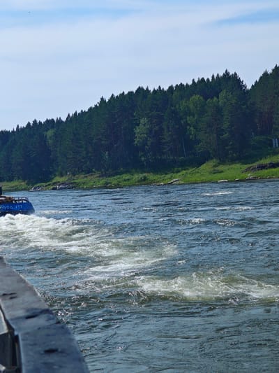 The image depicts a wide, turbulent river with a densely forested bank on the opposite side. In the foreground, a dark, weathered railing runs along the bottom edge of the frame, partially obscuring the view of the water. A blue boat, identified by Cyrillic lettering that reads "ВОДН0 717" (VodnO 717), is visible on the left side of the image, creating a wake of white foam as it moves through the water. The river itself is a deep blue, with ripples and churning white water indicating a strong current. The forest on the far bank consists of numerous evergreen trees, densely packed together, with a strip of green grass at the water's edge. The sky above is a clear blue with a few wispy white clouds. The scene appears to be a natural landscape with a functional boat operating on a river. The location is identified as Porog, Russia.