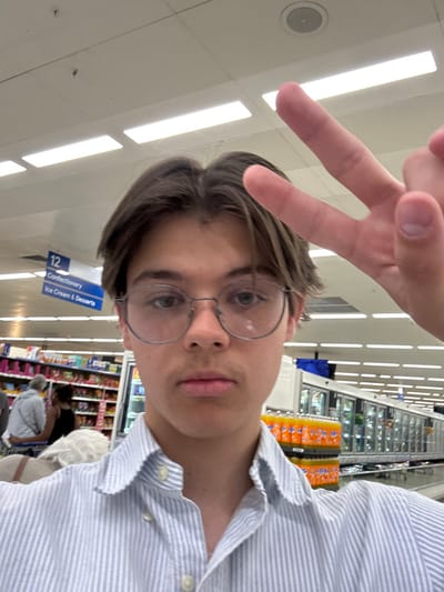 A young man with brown hair and glasses poses for a selfie in a brightly lit grocery store. He is wearing a striped blue shirt and making a peace sign with his hand. Shelves stocked with groceries are visible in the background, along with other shoppers. A sign for "Confectionery, Ice Cream & Desserts" can be seen near the man's head. The store lights are bright and run parallel to the aisles.