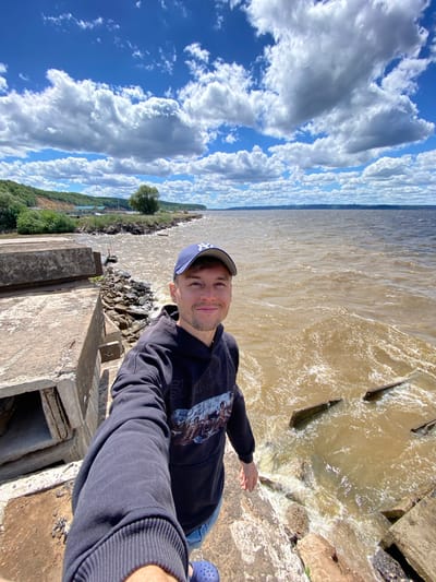 A man is taking a selfie on a sunny day in Leninsky, Russia, by a body of water. The man is wearing a dark hoodie with a graphic print and a New York Yankees baseball cap. He is standing on a concrete structure, which appears to be part of a pier or a seawall that is in disrepair. The water is a murky brown color and looks somewhat choppy, with some debris visible in the water, possibly parts of the ruined structure. In the background, there is a shoreline with green trees and a blue sky filled with white, puffy clouds. The overall impression is of an outdoor, casual setting on a warm day, with a focus on the man and the surrounding natural and man-made environment.