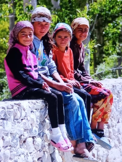 Four young females are seated side-by-side on a low, irregularly constructed stone wall in Kerayit, Uzbekistan. The wall is composed of light-colored, dry-stacked stones. Dense green foliage is visible in the background under bright daylight.

From left to right:
1.  A female, appearing adolescent, wears a purple and pink patterned headscarf, a pink and dark-striped long-sleeved top, black leggings, white socks, and pink slide sandals. She is looking forward with a slight smile.
2.  A female, appearing adolescent, wears a white and brown patterned headscarf, a light gray hooded sweatshirt with graphic elements, blue jeans with embroidered side patterns, white socks, and light-colored flip-flops. Dark hair is visible beneath her scarf. She is looking forward with a neutral expression.
3.  A female, appearing adolescent, wears a light blue and pink patterned headscarf, an orange long-sleeved shirt, and blue jeans. She is looking forward with a slight smile; her footwear is mostly obscured.
4.  A female, appearing adolescent, wears a beige patterned headscarf, a dark brown long-sleeved top, a multi-colored patterned skirt (red, orange, white), black socks with white patterns, and light blue flip-flops. She is looking forward with a slight smile.

All individuals have their hands resting in their laps.