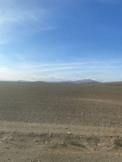The image presents a broad, open landscape in Jbilate الجبيلات, Morocco, under a clear blue sky. The sky dominates the upper portion of the frame, featuring subtle, elongated wisps of white clouds. Along the horizon, a distant range of mountains or hills appears, softened by haze.
The lower section of the image shows dry, barren ground, possibly agricultural land or a rocky plain. The terrain is textured with visible dirt and small stones, giving it a muted brown and gray appearance. Towards the right side of this foreground, two thin, light-colored posts stand upright, suggesting markers or parts of a boundary.
The scene captures a bright daytime with ample natural light, indicating fair weather. No individuals or specific activities are visible. The overall impression is one of vastness and solitude within an arid or semi-arid environment. There is no visible text in the image.