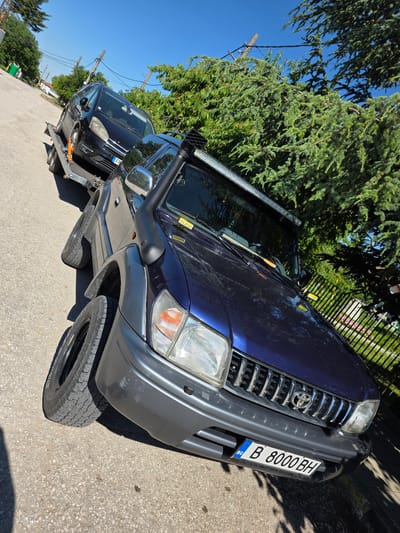 In Kalimantsi, Bulgaria, on a sunny day, a dark blue Toyota Land Cruiser Prado is parked on a paved road, with its front facing slightly towards the viewer's left. The vehicle is equipped with a snorkel and a light bar mounted on its roof. Behind it, a car trailer is attached, carrying a black minivan. The surrounding environment includes a paved road, a grassy area with a metal fence, and lush green trees. The sky is clear blue, indicating fair weather. The license plate of the Toyota is visible and reads "B 8000 BH".