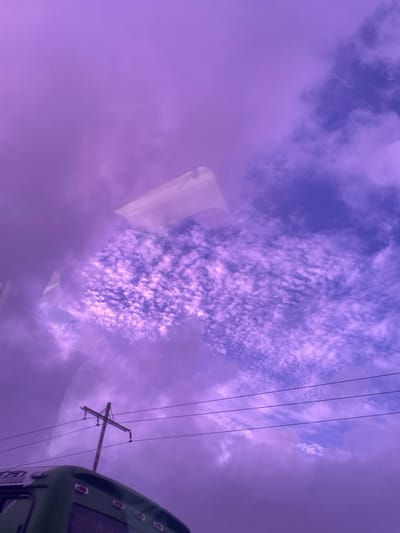 The image depicts an outdoor scene in La Cruz del Pastel, Venezuela, characterized by a predominantly purple and blue sky. The upper two-thirds of the frame are filled with clouds; dense, diffuse purple clouds occupy the left and upper sections, while a patch of textured white and purple altocumulus-like clouds is visible against a bluer sky background in the mid-right. A pale, indistinct, trapezoidal reflection, consistent with a building or structure, appears against the clouds in the upper-center, suggesting the image was captured through a reflective surface such as a window. In the lower-left quadrant, a light-colored wooden utility pole with a single crossarm is visible. Three dark utility wires extend horizontally from this pole across the lower portion of the frame. The upper-rear section of a dark green vehicle, possibly a bus, is partially visible along the bottom edge of the image, showing a curved roofline and a rectangular rear window. No individuals, specific actions, or direct interactions are visible.