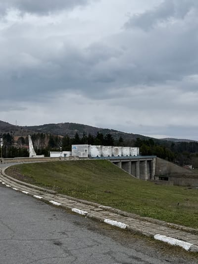 The image displays an overcast daytime scene of the dam and reservoir at Баня, Bulgaria, specifically the water management facility. In the foreground, a paved asphalt road curves slightly to the left, bordered by a concrete curb painted with alternating white and unpainted segments, indicating wear. To the right of the road, a grassy earthen embankment slopes upward towards a concrete dam structure.
The dam itself features a series of six vertical concrete supports visible below a blue railing. Above the railing, a row of six white, curved or conical structures, possibly sluice gates or spillway elements, are visible. To the left of these structures, a flat-roofed, white concrete building with blue graffiti or markings is situated. Further left, an additional smaller white building is present. Adjacent to the leftmost concrete dam structure, a tall, abstract, white monument-like structure, tapering upwards, stands on a raised platform.
Beyond the dam, dense green foliage and trees are visible, extending into forested hills that dominate the background. The sky is entirely covered with thick, dark grey clouds, suggesting an overcast day. No individuals or active interactions are visible within the frame.