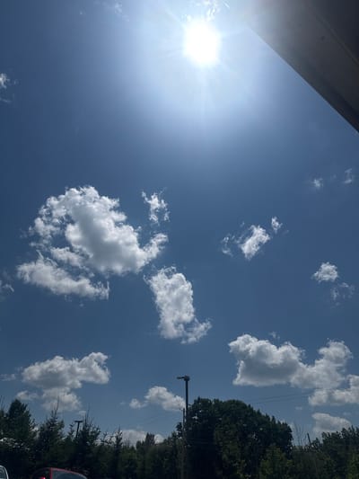 The image captures a bright, sunny day with a clear blue sky. The sun is visible in the upper portion of the frame, emitting strong rays. Several white, fluffy cumulus clouds are scattered across the sky. In the lower part of the image, the tops of dark green trees and a portion of a building's overhang are visible. A streetlight and power lines can also be seen above the trees. The scene appears to be an outdoor environment within the Village of Johnson City, United States of America, likely a public space or residential area given the presence of trees and infrastructure. There are no people or animals clearly depicted in the image.