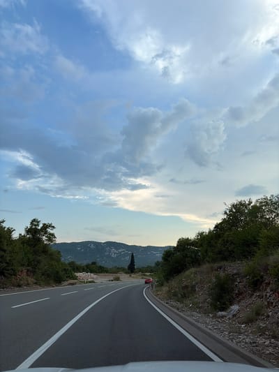 The image shows a curved asphalt road in Liješnje, Montenegro. The road has white lane markings and a curb. On the left side of the road, there are trees and dense vegetation. On the right side, there is a rocky embankment covered with grass and shrubs. In the distance, there are rolling hills covered with trees and some scattered buildings. A red car is visible on the road in the middle distance. The sky is filled with dramatic clouds, suggesting a potentially overcast or approaching storm. The overall scene depicts a rural or semi-rural setting in Montenegro, likely captured from a vehicle moving along the road.