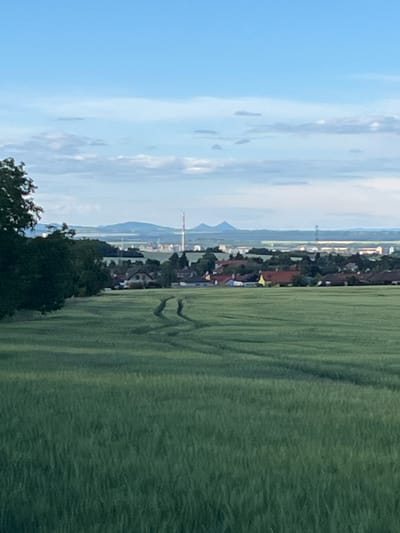 The image depicts a landscape view of Bašť, Czechia, on a partly cloudy day. In the foreground, a vast green field dominates the scene, with visible tracks from a vehicle creating a winding pattern. Beyond the field, a cluster of houses with red and brown roofs forms a village. Further in the distance, a city skyline is visible, characterized by buildings and a tall, slender red and white tower. The horizon is marked by rolling hills and mountains under a pale blue sky with scattered clouds. A dense dark green tree occupies the left side of the frame, partially obscuring the view of the village and distant city.