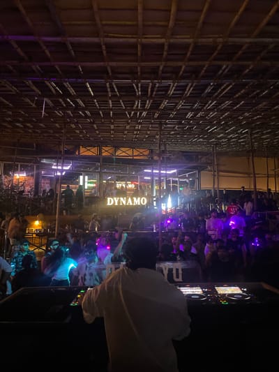The image depicts a nighttime scene at a crowded open-air venue, likely a nightclub or bar, with a bamboo-like ceiling structure. A DJ booth is prominently featured in the foreground, with a person in a white shirt facing away from the camera, operating the equipment. The crowd is packed together, with many individuals dancing and some raising their hands. Neon lights, predominantly purple and blue, illuminate the scene, creating a vibrant and energetic atmosphere. Behind the crowd, a building with a glass facade is visible, featuring illuminated signage that reads "DYNAMO" in large, bright letters. This suggests the venue is named Dynamo. The overall impression is that of a lively party or event taking place in Vagator, India, known for its nightlife.