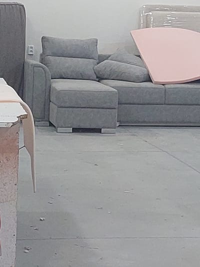 The image shows a close-up of a gray sectional sofa with plush cushions and a light pink curved accent piece. The sofa appears to be in a room with a light-colored wall and a gray concrete wall in the background. There is an electrical outlet on the wall near the sofa. In the foreground, part of another piece of furniture, possibly a wooden bench or a low table, is visible, along with some construction materials. The scene is brightly lit, suggesting it is daytime. There is no clear indication of any activity or people in the image. The overall impression is that of a room being furnished or redecorated, or perhaps a storage area for furniture. The location is identified as Dalar, Armenia.
