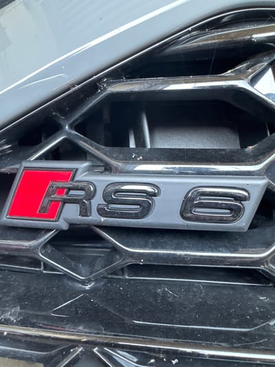 This is a close-up, eye-level shot of the "RS6" emblem on the front grille of a car. The emblem is vertically oriented and centrally located. It has a matte gray backing with glossy black lettering. The top of the emblem features the letters "R" and "S," while the bottom displays the number "6" over a bright red rectangular section with the "6" design continuing in black. The grille itself is a glossy black, diamond-patterned structure. The surrounding car body, visible on the left, is a light gray color. The lighting suggests it is daytime, and there are no prominent weather cues. The image appears to be taken in a natural or outdoor setting, possibly a driveway or parking area in Wettstetten, Germany, given the location context. No people are visible. The focus is solely on the car's emblem and grille, highlighting its design and branding. There is some dust and minor scratches visible on the grille and emblem, suggesting the vehicle is in use.
