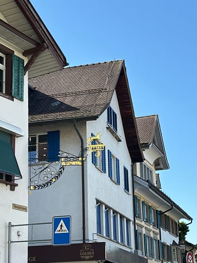 The image shows a street scene in Wald (ZH), Switzerland, with buildings and a clear blue sky. In the foreground, a white building has a dark brown roofline with visible wooden support beams. To its right, a lighter colored building with a brown tiled roof features blue shutters on its windows. A decorative gold sign depicting a horse is mounted on the wall of this building, above a black wrought-iron scrollwork element. A street sign that reads "Tössalstrasse" is visible on the white building to the left. Below this, a blue and white pedestrian crossing sign is mounted on a metal pole. Further to the right, a series of buildings with various window styles and shutters are visible, suggesting a European architectural style. The overall scene depicts a quiet, sunlit street.