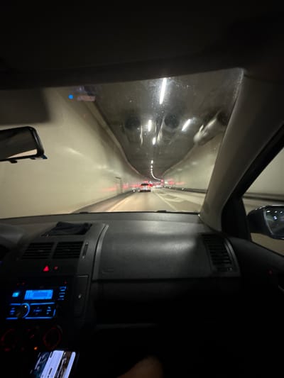 The image depicts a view from inside a vehicle, possibly a truck, traveling through a tunnel. The tunnel is illuminated by overhead lights, and other vehicles are visible ahead in the distance. The vehicle's dashboard is visible on the right side of the frame, showing various controls and a display screen. Based on the presence of a tunnel and the city context, it's plausible the location is near Liestal, Switzerland, possibly on the A22 motorway which contains a tunnel near Liestal. However, positive identification requires location specific details from the picture.