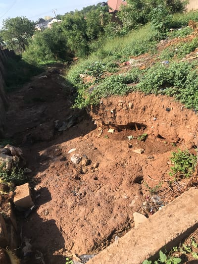 This media file captures an outdoor scene dominated by earthy tones and green vegetation, taken during what appears to be a bright, sunny day. The image is oriented vertically (or rotated 90 degrees clockwise).
The foreground and midground feature a significant amount of reddish-brown dirt, indicating an unpaved or excavated area. A noticeable depression or gully runs through the dirt, suggesting either a natural erosion channel or a man-made ditch. Scattered within the dirt and along the edges of the channel are various pieces of refuse, including plastic bottles, wrappers, and general litter, indicating an issue with waste disposal.
Lush green foliage and shrubs densely cover the left side of the image, with some patches appearing wild and overgrown. There are also dried branches and sparse vegetation closer to the dirt path. A sturdy, light-colored concrete slab or structure is partially visible on the far right, bordering the dirt area.
In the background, on the far left, several residential buildings with distinct rooftops can be seen nestled among trees. Utility poles and wires are also discernible against a clear, light blue sky, hinting at a developed, possibly suburban or semi-urban setting. The overall impression is that of a neglected or undeveloped area adjacent to residential zones. No people are visible, and there is no discernible activity taking place. There is no visible text in the image. The sunlight casts distinct shadows, particularly from the concrete structure and some vegetation, confirming it is daytime.