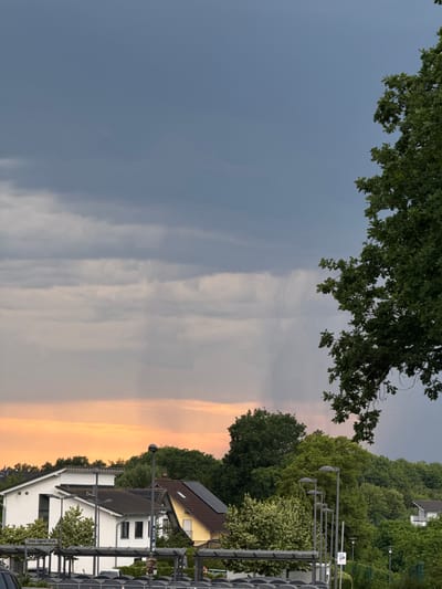 The image captures a view of Sankt Augustin, Germany, featuring a sky with dramatic clouds, some of which are dark and ominous, suggesting an impending rain shower. A visible rain shaft is descending from the clouds in the mid-ground. The foreground displays residential buildings with white and yellow facades, some equipped with solar panels. Lush green trees frame the scene, with a large tree on the right side of the frame dominating the upper portion of the image. A covered structure, likely a bus stop or parking shelter, is present in the lower part of the frame, with lampposts visible nearby. The overall atmosphere is one of changing weather conditions in a suburban setting.