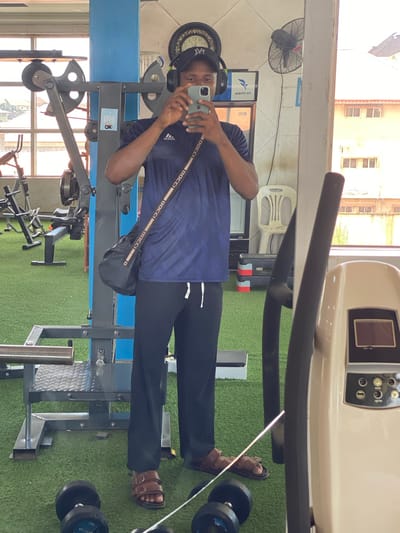 This media file shows a man in a gym in Podo, Nigeria. The man is taking a selfie in a mirror, and he is wearing headphones, a blue t-shirt, black pants, and brown sandals. He has a black bag slung across his chest. In the reflection, we can see various gym equipment, including weight machines, dumbbells, and a treadmill. A fan is mounted on the wall, and a clock is visible above it. The setting appears to be a well-lit indoor gym with green turf flooring. The time of day is not explicitly stated, but the lighting suggests it could be daytime. The overall mood seems casual and relaxed, as the man is engaged in a personal activity within the gym environment. Visible text includes "BOCCHI" on the man's bag, and some text on the gym equipment and the fan. The overall impression is of a typical gym scene with a focus on the individual's presence.