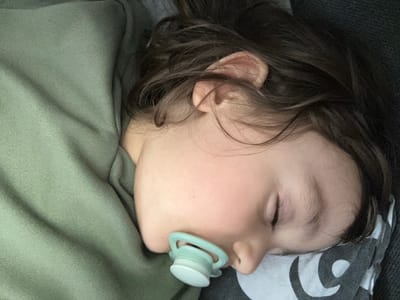 A young child is depicted sleeping on their right side, with their eyes closed. A light mint green and white pacifier is in the child's mouth. The child has dark brown, slightly wavy hair fanned around their head and ear. They are wearing a long-sleeved top made of a ribbed, muted olive green fabric. The child's head rests on a pillow featuring a gray and white abstract or patterned design. The location is in the city of Fulda, Germany.