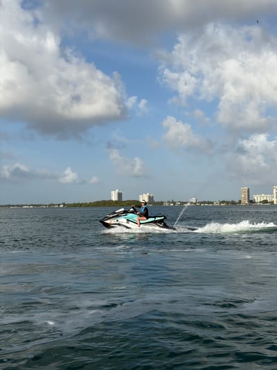 The image depicts a jet ski operating in the waters off the coast of North Miami, Florida. A person is riding the jet ski, which is creating a spray of water as it moves. The shoreline of North Miami is visible in the background with buildings. The sky is partly cloudy with patches of blue. The water appears choppy.