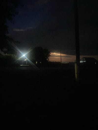 The image shows a nighttime scene in a rural area, likely in Жданов ауылдық округі, Kazakhstan. The photograph is vertically oriented and appears to be taken from a low angle, looking upwards. The primary subject is a bright, starburst-shaped light source, likely a lamp or streetlight, positioned in the mid-ground. This light illuminates the surrounding area, creating strong lens flares.
In the background, a faint horizon line is visible, with hints of clouds and possibly distant lights. A dark, vertical pole runs across the upper left portion of the frame, suggesting the presence of a power line or similar structure. Silhouetted trees and foliage are visible in the foreground and to the left of the light source, indicating an outdoor, natural setting. The overall impression is one of a quiet, possibly somewhat isolated, rural night. The lighting suggests it is nighttime, and the presence of clouds might imply overcast or potentially stormy weather, though this is not definitively clear. There is no visible text in the image.