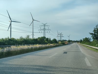 The image shows a highway in Damlos, Germany, with wind turbines and power lines visible on the left side of the road. The sky is a clear blue with some scattered clouds. The road is asphalt and has white lines marking the lanes. There is a guardrail on the right side of the road, and a grassy embankment with tall dry grass on the left. In the distance, there are trees and fields. The scene appears to be a sunny day.