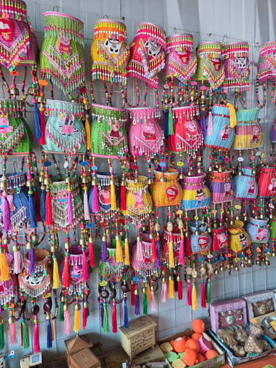 This image captures a vibrant display of numerous colorful, handcrafted woven items, likely baskets or decorative lanterns, arranged in rows on a white grid structure. The scene suggests a retail environment, such as a market stall or shop, in Jiejinkou, China.

The main subjects are small, brightly colored baskets, predominantly made from woven plastic or raffia in shades of pink, green, blue, yellow, red, and purple. Many are two-toned and feature cartoon characters, most notably Peppa Pig in various poses. Other baskets display stylized human-like figures or unique animal characters. Each item is extensively decorated with long strings of small, multicolored beads that form intricate patterns, often interspersed with larger wooden beads and small brass bells. Vibrant tassels in a wide array of colors—red, pink, yellow, blue, green, and purple—hang from the bottom of many of the baskets. Some decorations include small plastic charms.

To the right, on shelves, additional goods are visible. These include boxes labeled "Happy Porcelain Tea Set" and "Mari Porcelain Tea Set," depicting white animal-shaped teacups. Also present are piles of bright orange, fruit-shaped plastic toys, and other packaged items with visible text like "POPINY," "P-UP," and "FELI." Small yellow price tags or product labels are attached to some of the hanging decorations.

The activity depicted is the display of goods for sale. The overall impression is one of cheerfulness and abundance, characteristic of a lively market. The setting is indoors, with no discernible time of day or specific weather conditions.