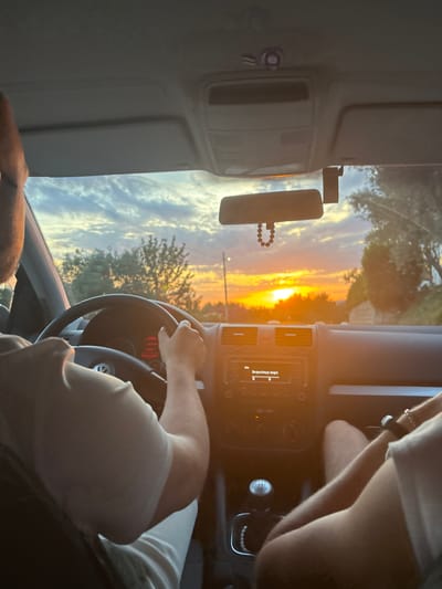 An interior shot of a vehicle, likely a Volkswagen due to the steering wheel emblem, captures a sunset scene in Gornji Katun, Montenegro. The driver, an adult male wearing a light-colored short-sleeved shirt, is positioned to the left, with his right hand on the steering wheel. A passenger, also an adult wearing a similar light-colored shirt and a wrist-worn accessory, is visible on the right, with their lower body and right arm in frame.
The car's interior features a dark gray dashboard, a manual gear shift with a 5-speed pattern, and an infotainment system displaying "Info" and "Bespechnyy angel" (Carefree angel). A rearview mirror, adorned with a beaded rosary-like object, hangs from the ceiling, which is light gray. A black dashcam or similar device with a blue indicator light is attached to the windshield to the right of the rearview mirror.
Outside the vehicle, a prominent golden-orange sun is setting on the horizon, casting a strong glare and illuminating the sky with vibrant orange, yellow, and light blue hues. Scattered clouds are visible against the colorful sky. Silhouetted trees line the road ahead, and a utility pole or thin vertical structure is discernible on the left side of the frame.