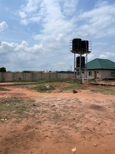 The image shows an outdoor scene with a focus on a water tower and a building. The ground is covered in red earth and patches of green grass, with some scattered debris. In the background, a concrete wall with green posts separates the property from a street with more buildings and utility poles. A single-story building with a green, gabled roof is visible on the right, and a metal water tower with two large black water tanks stands prominently to the left of the building. The sky is partly cloudy with a light blue hue. There are no people or animals visible in the scene. The scene appears to be in a developing area, possibly within the city of Ekodel, Nigeria, given the architectural style of the building and the presence of overhead utility lines.