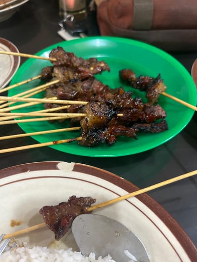 In this close-up shot, skewers of grilled meat are presented on two plates. The foreground features a green plate filled with several satay skewers, the meat appearing dark and well-cooked with some charred edges. The skewers are made of light-colored wood. To the right, a larger, off-white plate with a brown rim holds a single satay skewer alongside a serving of white rice. A silver spoon is partially visible, resting on the rice. The lighting suggests an indoor setting, possibly a restaurant, with a focus on the food. The image captures a meal, likely satay, being served. The composition is tight, emphasizing the textures and colors of the grilled meat and rice. No people or specific environmental details beyond the food and serving ware are clearly visible. The context of Cotpuuk, Indonesia, implies the cultural significance of this dish.