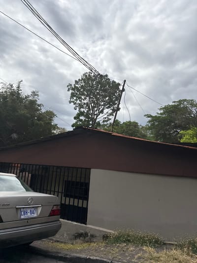 The image shows a residential building in Escazú, Costa Rica, located at coordinates 9.924 latitude and -84.142 longitude. The building is a single-story, two-toned structure with a brown roof and white walls. It has a barred window and a silver car parked in front of it. The car's license plate reads "CBH-847." Several power lines run across the sky, which is overcast with gray clouds. A medium-sized tree grows on the roof of the building, and other trees line the street. The scene appears to be on a residential street, with no people or animals visible.