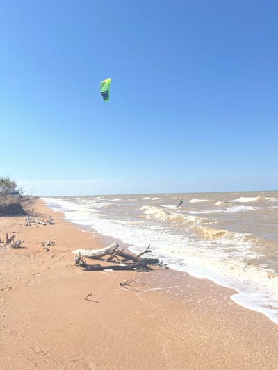 The image depicts a sandy beach on a clear, sunny day. In the distance, a kitesurfer is visible riding waves in the sea. A large, green kite is also visible in the sky. The foreground of the beach is scattered with driftwood and shells. The overall scene suggests a recreational activity taking place in a coastal environment. The location is identified as Dolzhanskaya, Russia, a popular resort town known for its beaches and water sports.