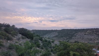 The image depicts a natural landscape with rolling hills covered in dense vegetation. The sky is overcast with hints of pastel colors, suggesting it might be dawn or dusk. The terrain is rugged, with rocky outcrops and patches of dry grass visible on the slopes. Several trees, some with full foliage and others appearing more sparse, dot the landscape, particularly in the foreground and along the hillsides. In the far right of the image, a few white vans and what appears to be a building with a red roof are visible, hinting at human presence and infrastructure in the vicinity. The overall scene conveys a sense of rural tranquility and natural beauty.